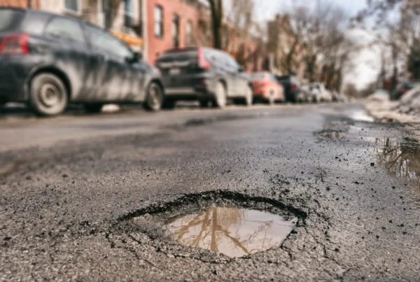 A large water-filled pothole in a cracked asphalt street with cars parked along the curb in the background.