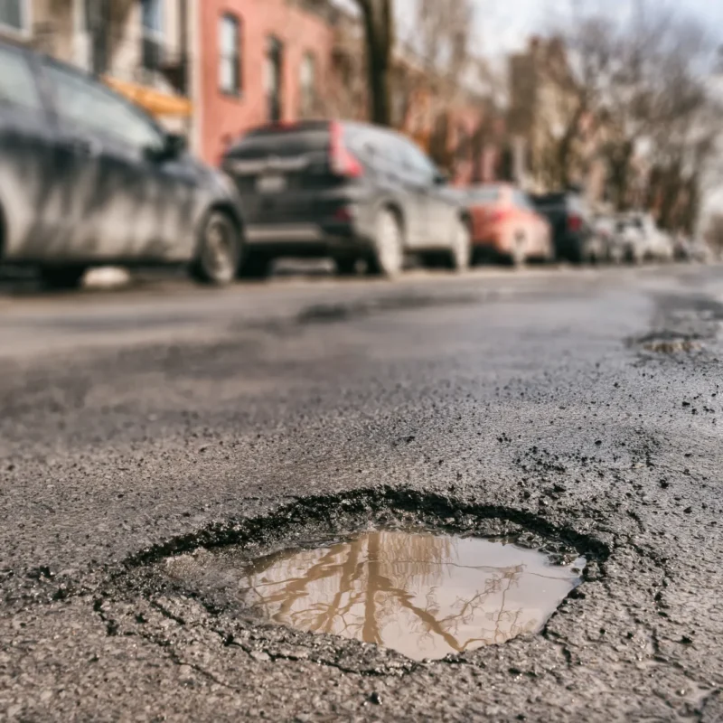 A large water-filled pothole in a cracked asphalt street with cars parked along the curb in the background.