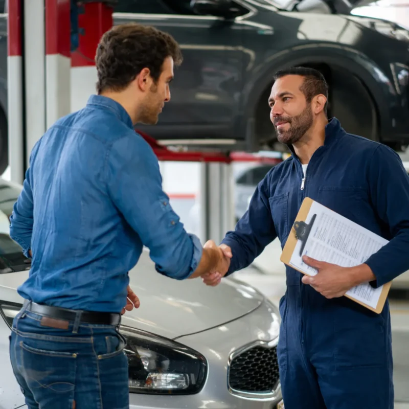 A mechanic in a navy coverall holding a clipboard shakes hands with a customer in an auto repair shop, with a car beside them and an SUV on a lift behind.