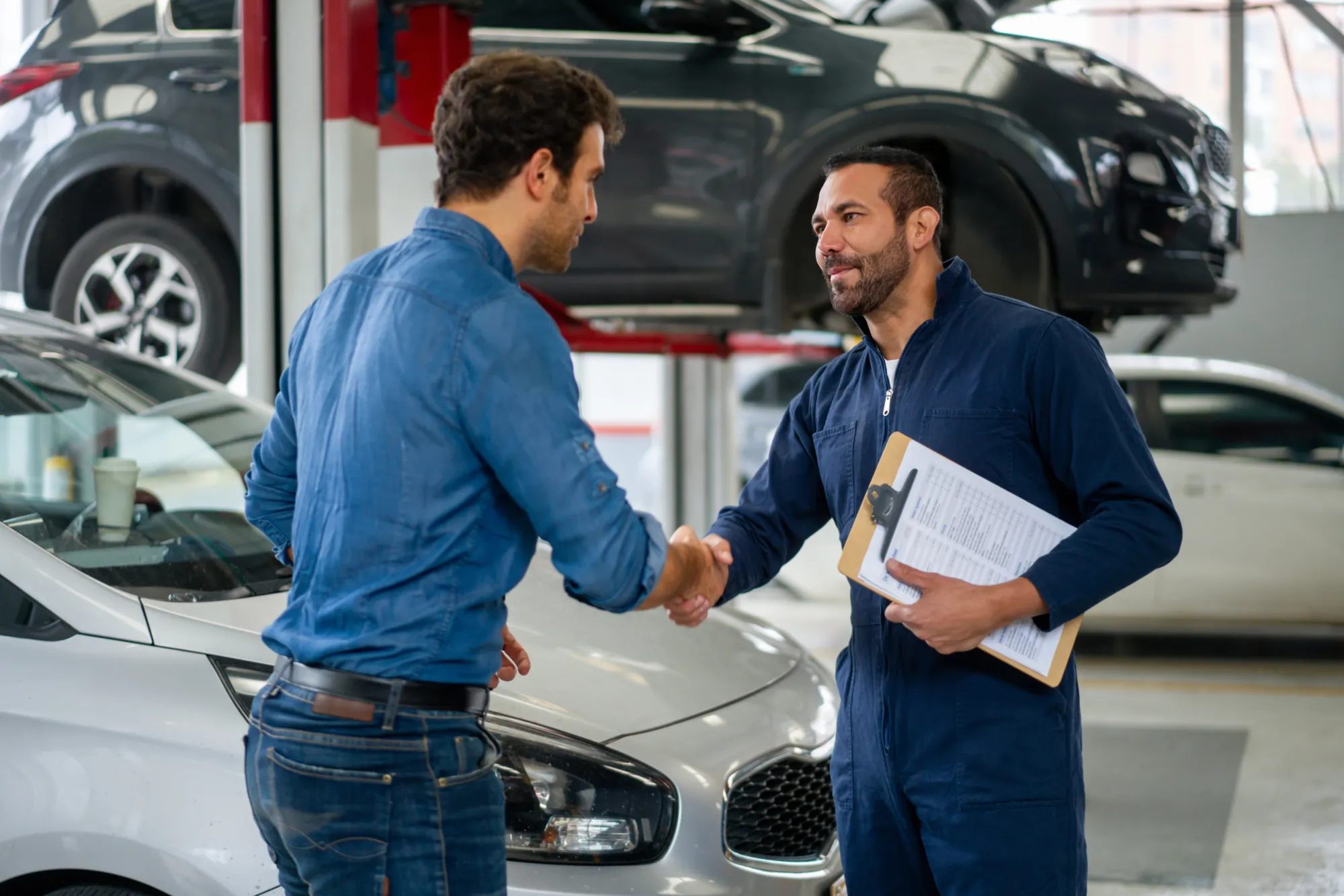A mechanic in a navy coverall holding a clipboard shakes hands with a customer in an auto repair shop, with a car beside them and an SUV on a lift behind.