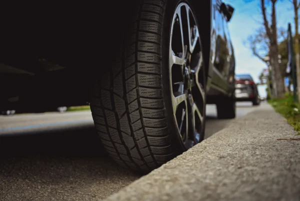 Close-up low-angle view of a car's black tire and alloy wheel parked next to a concrete curb on a tree-lined street.
