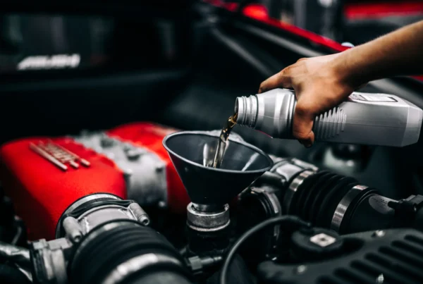 A hand pours motor oil from a bottle through a black funnel into the filler neck of a car engine with a red intake cover.