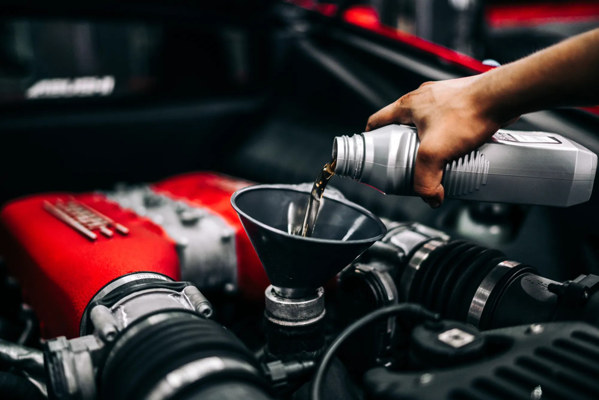 A hand pours motor oil from a bottle through a black funnel into the filler neck of a car engine with a red intake cover.